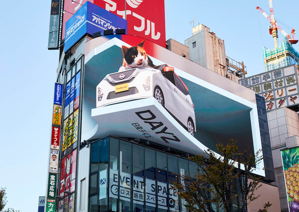 ねこ用軽自動車「にゃっさんデイズ」に乗って舌なめずりをする巨大猫 by クロス新宿の街頭ビジョン